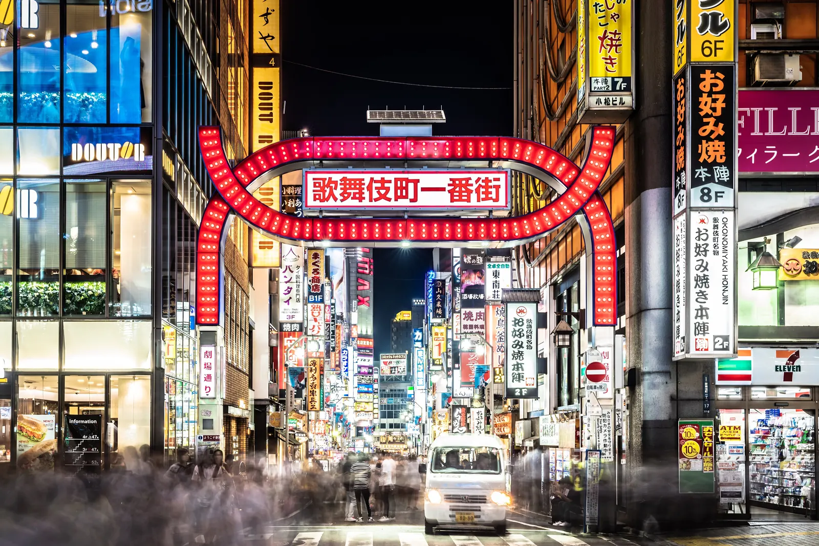 Das rote, nachtbeleuchtete Bogentor Kabukichō Ichibangai, mit Neonschildern entlang der Straße dahinter und einem weißen Van an der Kreuzung