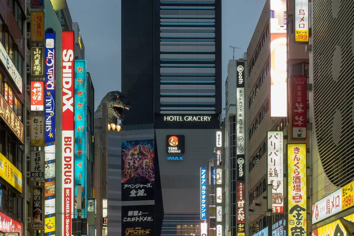 En smal gade i Shinjuku med lodrette skilte i tusmørket og et Godzilla-hoved, der kigger ud over tagene ved siden af et højt hotelttårn