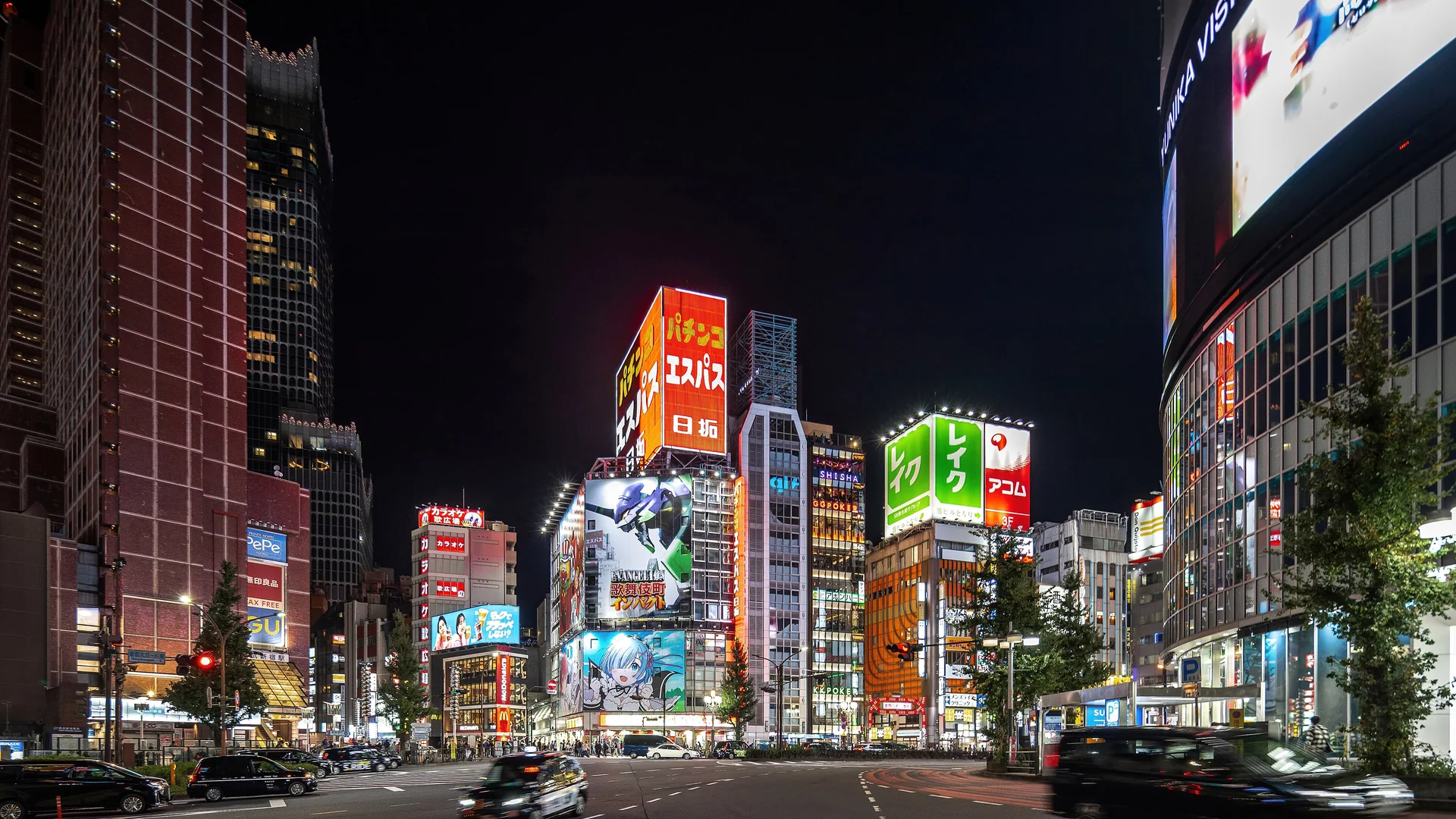 Illuminierte Türme mit roten, orangefarbenen und grünen Leuchtreklamen ragen in einen pechschwarzen Nachthimmel über einer breiten Shinjuku-Kreuzung, auf der Autos vorbeifahren; ein Bild des Viertels ohne Bezug zur Aufführung