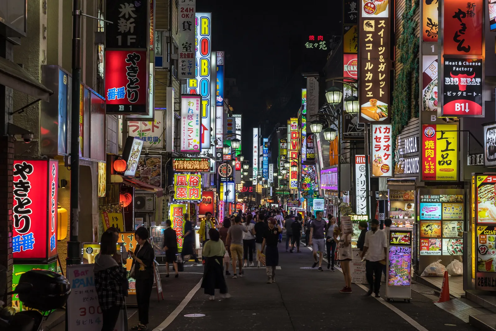 Eine schmale Straße in Kabukichō bei Nacht, gesäumt von beleuchteten vertikalen Schildern und Menschenmengen in der Ferne